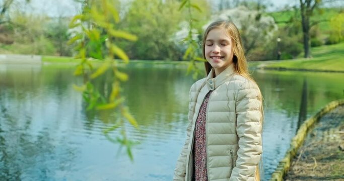 Smiling preteen girl standing near pond in the park and smiling at camera. Girl has long blond hair