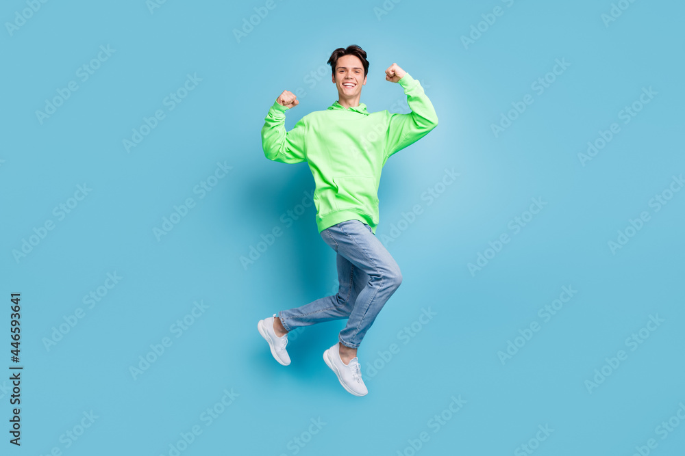 Wall mural Photo of cheerful strong muscular guy jump show biceps wear green hoodie jeans shoes isolated blue color background