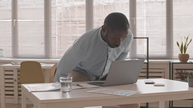 Arc of young African man wearing formal shirt, sitting down at desk in office at daytime, starting typing on portable computer