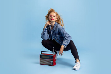 Portrait of attractive young girl in retro 90s fashion style, outfits posing isolated over blue studio background. Concept of eras comparison, beauty, fashion and youth.
