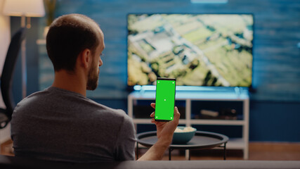 Caucasian man vertically holding smartphone with green screen display, looking at chroma key background for isolated blank design used for digital mockup template. Copy space in living room