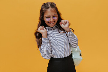 Excited teenager rejoices on orange background. Tanned happy girl in school uniform smiles and holds backpack.