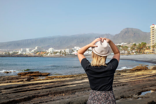 Rear view of young Caucasian woman holding her hat with both hands and looking towards the entire coast of Las Americas, Costa Adeje, with its popular tourist resorts, Tenerife, Canary Islands, Spain