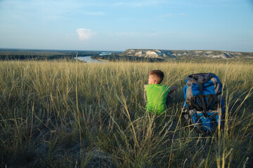 A boy in a green T-shirt sits next to a tourist backpack rests and looks into the distance, among the wild meadow grass against the backdrop of wild nature on a journey