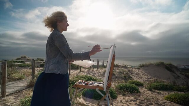 Middle-aged European woman artist paints a picture with paints overlooking the sea. Blue skies. Close-up.