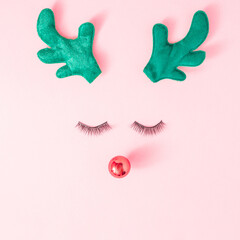 Christmas reindeer concept made of plush deer ears, red bauble decoration and artificial eyelashes on pastel pink background. Minimal winter holidays idea. Flat lay top view composition.