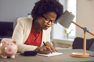 Positive millennial african american business woman carefully writes notes sitting near calculator and piggy bank. Woman checks the bills, keeps track of expenses and plans the budget. - obrazy, fototapety, plakaty