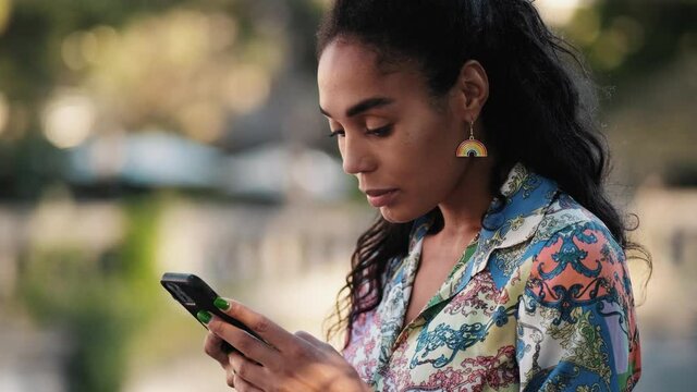 A happy african american woman with rainbow earrings is typing message using her phone during pride gay parade outside
