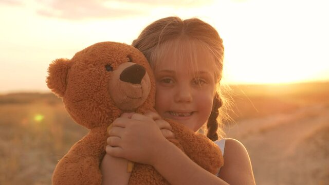Cute happy white girl hugs her favorite soft toy. A girl smiles at the camera at sunset in the park with a teddy bear in her hands. A child's favorite play with a teddy bear. Happy child smiles.