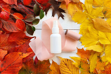 Unbranded white cosmetic spray bottle, silk pink ribbon and round frame of bright autumn or fall leaves on white wood background. Mockup. Skincare beauty and liquid antibacterial spray concept.