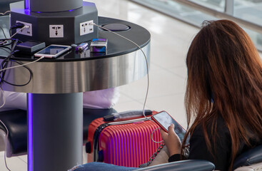 A young woman sitting at a charging station and looking at her smartphone. Recharging mobile phones from free charge station at the airport. - obrazy, fototapety, plakaty