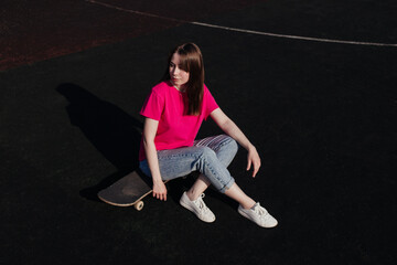 Young woman in pink t-shirt sitting on skateboard outside looking to the right. Summer activity, healthy lifestyle, workout, sport, fitness, riding. Female skater. Having fun outdoors.