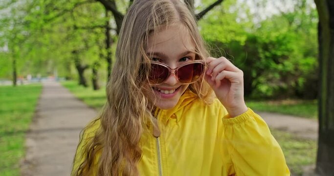 Cheerful preteen girl 10s with long blonde hair weared in yellow jacket in the city park. Child in sunglasses smiling at camera