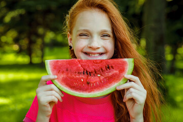 A beautiful young girl with red hair in a red dress holds a slice of juicy watermelon in nature on a warm summer day. Healthy lifestyle concept.