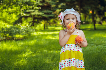 A beautiful little girl in a white panama hat and in a yellow dress eats a peach outdoors in a city park. Healthy lifestyle concept. Funny photo. Space for text.