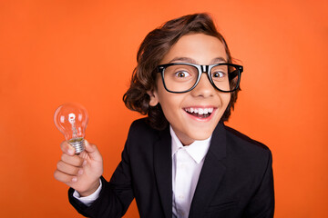 Photo of cheerful young amazed small boy hold light bulb idea plan smart isolated on orange color background