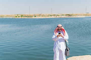 Saudi Arabian man, in traditional dress, standing by the sea, checking his mobile phone.