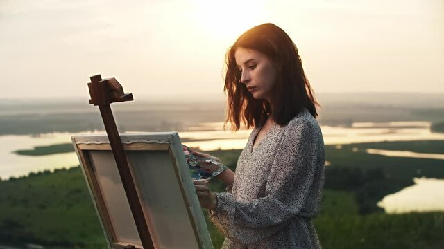 A young beautiful woman artist draws a painting on an easel at sunset field