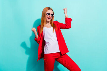 Photo of shiny lucky orange hair woman dressed red jacket dark eyewear rising fists smiling isolated cyan color background