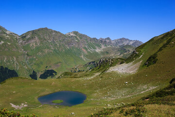 The Main Caucasian Ridge and blue mountain lake surrounded with grass and flowers of the alpine meadows