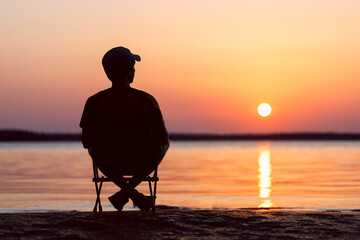 Man is sitting in a camping chair on the shore of the sunset sea. Background is blurred.