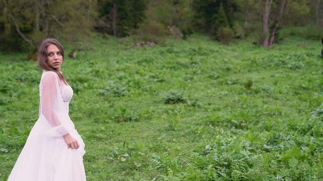 A young beautiful woman in a white wedding dress stands in a field with horses.