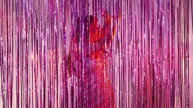 Birthday party. Happy glamorous woman. Holiday fun. Festive celebration. Joyful lady silver spangle dress dancing behind shimmering purple pink cascade curtain defocused copy space.