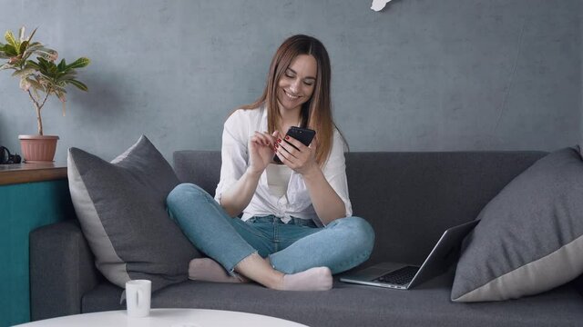 young woman using smart phone surfing social media, checking news, playing mobile games or texting messages sitting on sofa.