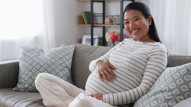 pregnancy, rest, people and expectation concept - happy smiling pregnant asian woman sitting on sofa at home