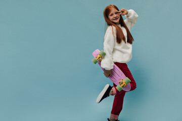 Beautiful girl with ginger hair in cute white sweater and red stylish trousers holding pink skateboard and peace sign on isolated backdrop..
