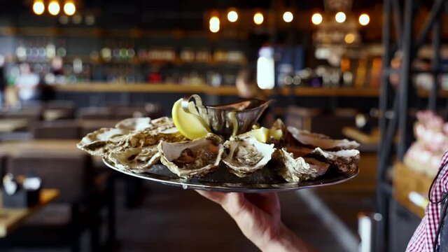 Waiter carries a tray of served oysters on ice with lemon for luxury restaurant visitors. Garcon brings salver large plate traditional meditarian dish sea food for gourmets.