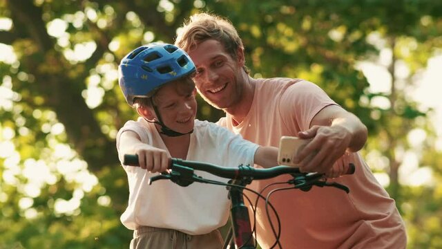 A smiling father is taking selfie photo with his young son while he is teaching to ride a bicycle outdoors in the park at summer