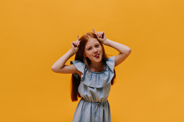 Happy pretty little funny girl grimaces. Cool redhead child in blue dress shows tongue and makes ears with her fingers on orange background.