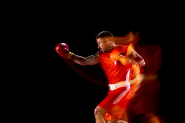 Young professional boxer in red shorts and gloves training, exercising over black background in...