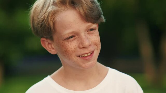 A happy boy with freckles is looking to the camera standing in the park at summer