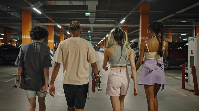 Back view of group of happy diverse friends walking together through underground parking zone, tracking shot