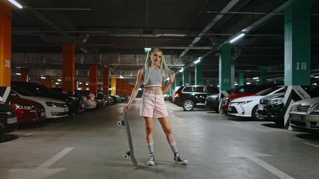 Young woman with bright unusual appearance posing with skateboard at underground parking zone, flirting to camera