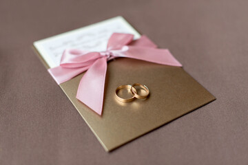 Close-up of two golden wedding rings on a invitation card. Wedding symbols, attributes. Golden wedding invitation card with pink bow on a brown background