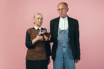 Serious elderly man and woman in retro vintage farmer outfits isolated on pink studio background. Retro style, comparison of eras and cultural concept.