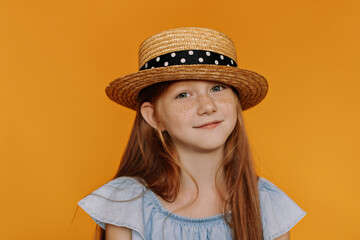 Portrait of charming little redhead girl with freckles smiling. Pretty child in boater and blue blouse looks into camera on orange background.