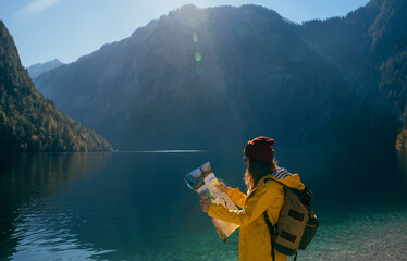 a girl in a yellow jacket and with a backpack in sunny weather looks at map at mountains near a blue lake side view