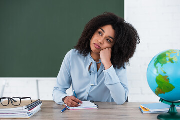 Pensive african american teacher sitting near stationery, eyeglasses and globe in classroom