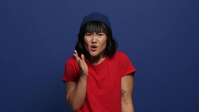 A shocked confused asian woman is looking to the camera isolated over blue wall in the studio
