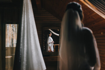 Obraz na płótnie Canvas A beautiful bride in lingerie with a long veil stands in front of a mirror and is reflected in it in a wooden room. Wedding photography.