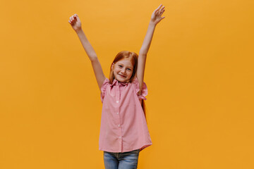 Redhead charming little girl rejoices. Pretty child with freckles dressed in striped shirt rises arms on isolated orange background.