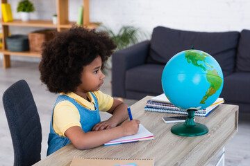 Side view of african american girl writing on notebook and looking at globe at home