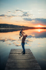 father raised his little son up standing on a wooden bridge against the background of a large lake and beautiful sunset in summer side view