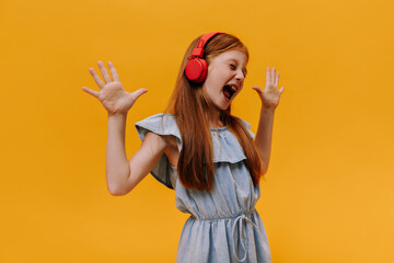 Emotional young redhead child in blue dress sings loudly, screams and listens to music in headphones on orange background.