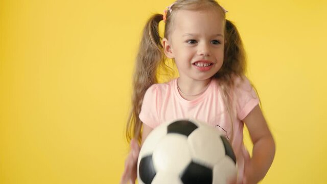 Authentic cute smiling preschool little girl with classic black and white soccer ball look at camera on yellow background. child play football in t-shirt and shorts. Sport, championship, team concept