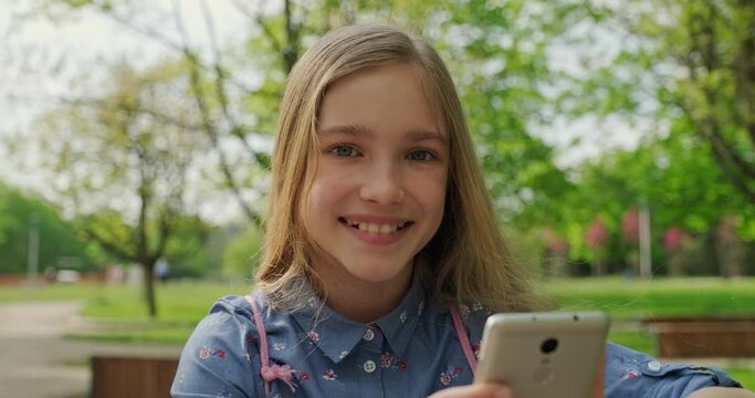 Preteen girl 10 years old sitting on the bench in the city park. Child smiling with teeth. Gimbal shot. Slow motion